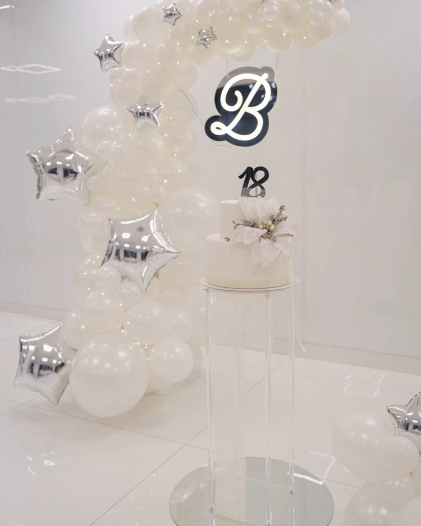 Arco di palloncini bianchi con stelle argento, iniziale luminosa e torta bianca su una colonna trasparente