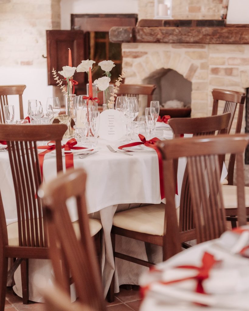 Sala con camino in pietra e tavoli rotondi apparecchiati con nastri rossi, rose bianche e candele rosse