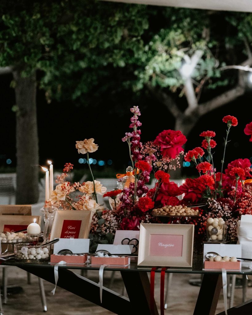 Confettata all’aperto di sera, tra fiori rossi e pesca, candele e cartellini dei confetti