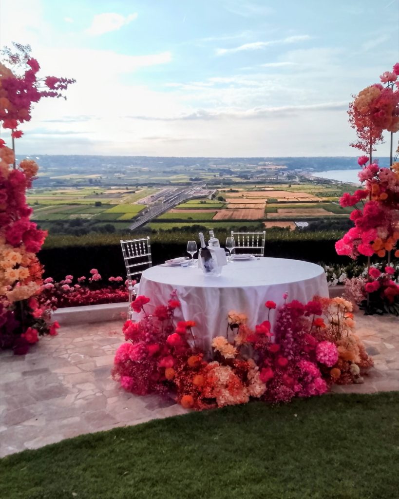 Tavolo degli sposi tra due colonne di fiori rossi, pesca e fucsia, con vista sulla campagna e sul mare